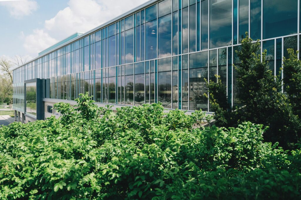 Contemporary building facade with glass panels reflecting nature, surrounded by lush greenery.