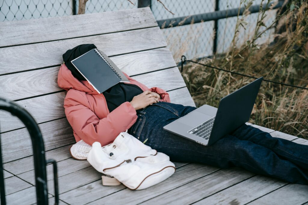 Exhausted student napping on a bench outdoors with a laptop and notepad on face.